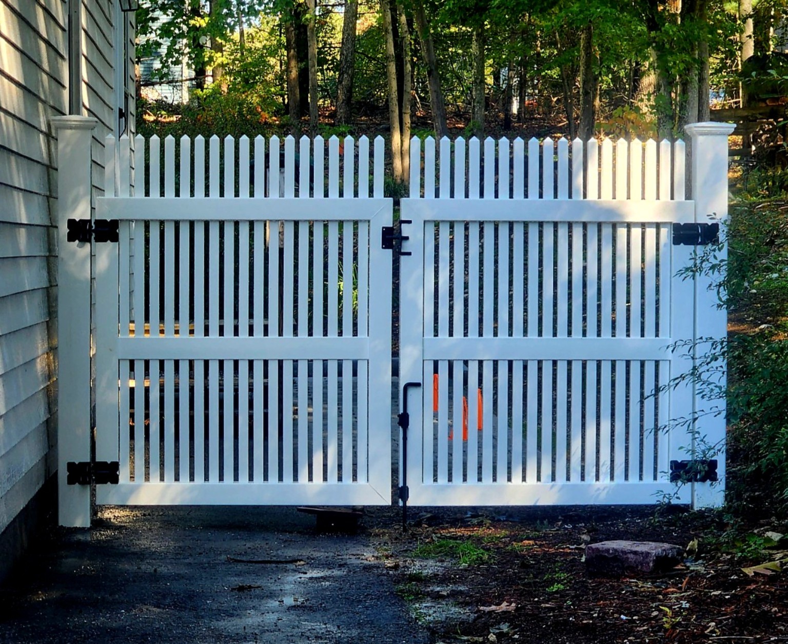 White vinyl double picket gate with black hinges and a center latch beside a house.